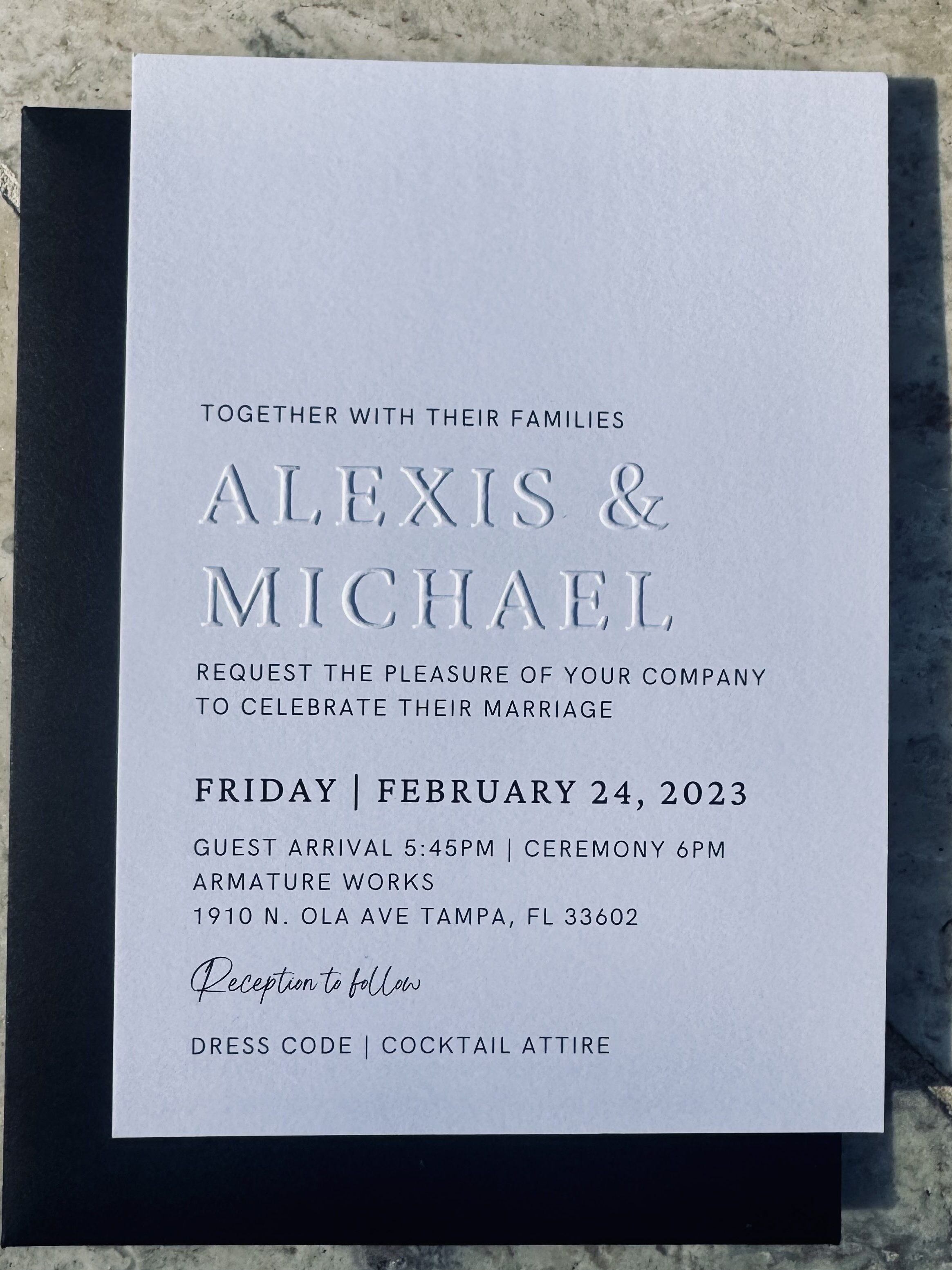 An elegant wedding invitation featuring raised lettering and embossed design on thick white paper, accompanied by a black backing.